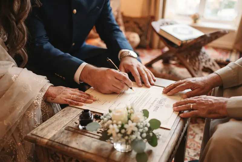 A bride and groom signing the Nikah contract at a low table, with a family elder beside them and a Quran on a stand behind